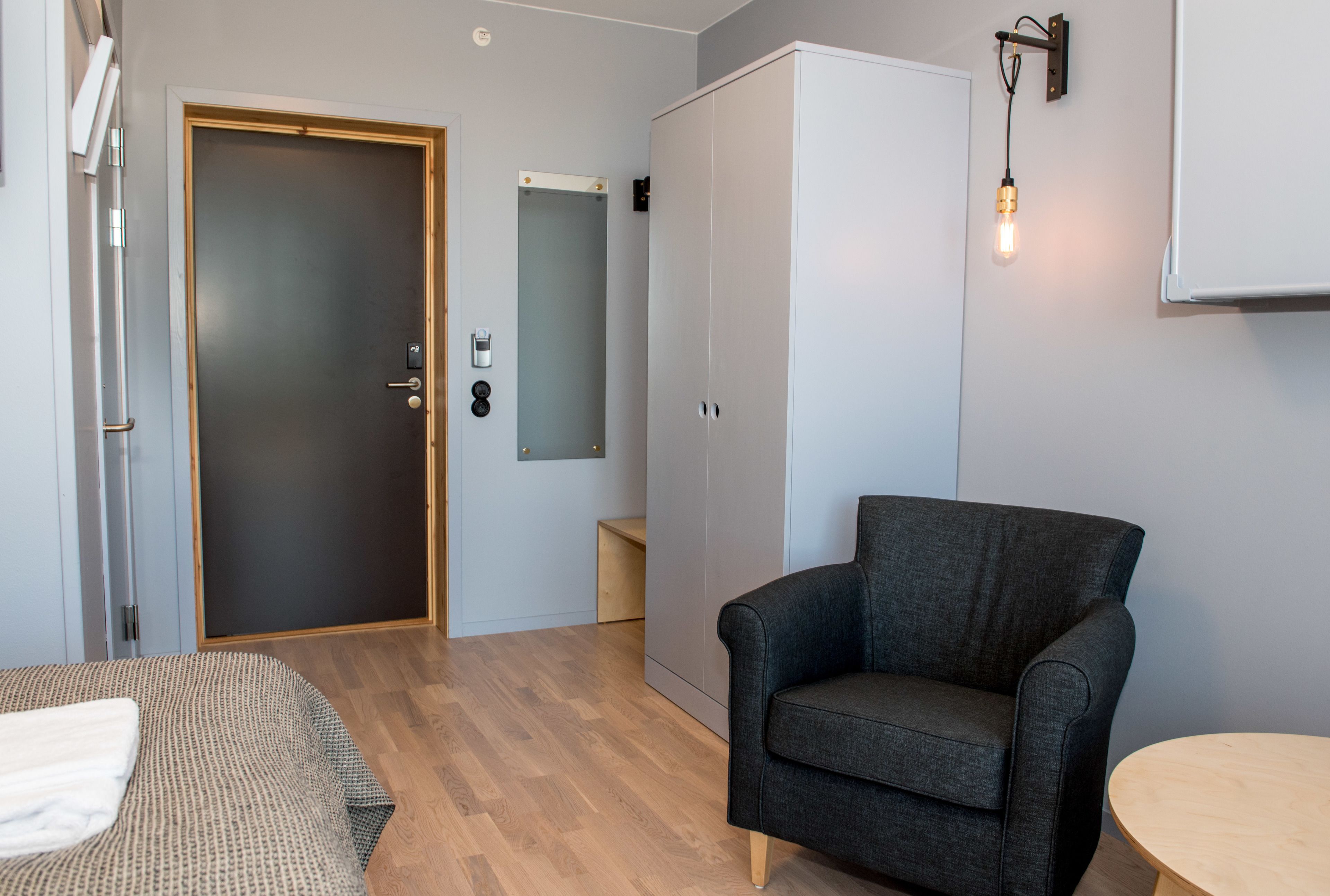 A room with a dark grey armchair, a wooden floor, a wardrobe, and a small round table.