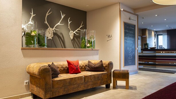 A modern living room with a brown leather sofa, a small wooden side table, and a wall-mounted shelf with decorative glass vases and antler sculptures. - Müllers Landhotel (Mespelbrunn)
