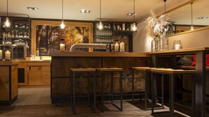 A modern bar area with wooden stools, hanging pendant lights, and a large mural. - Müllers Landhotel (Mespelbrunn)