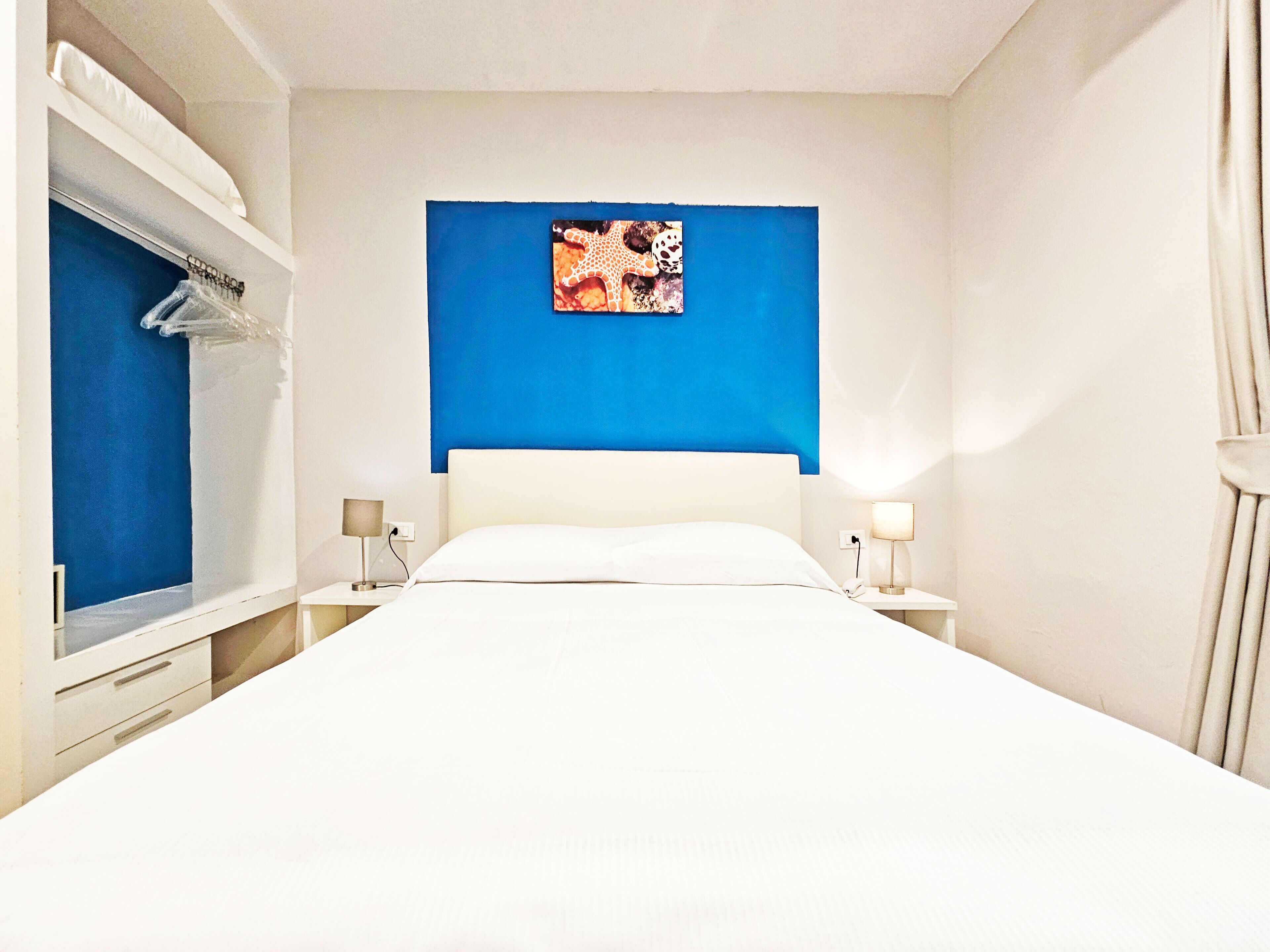 A hotel room with a bed, two bedside tables with lamps, a wardrobe, and a blue accent wall.