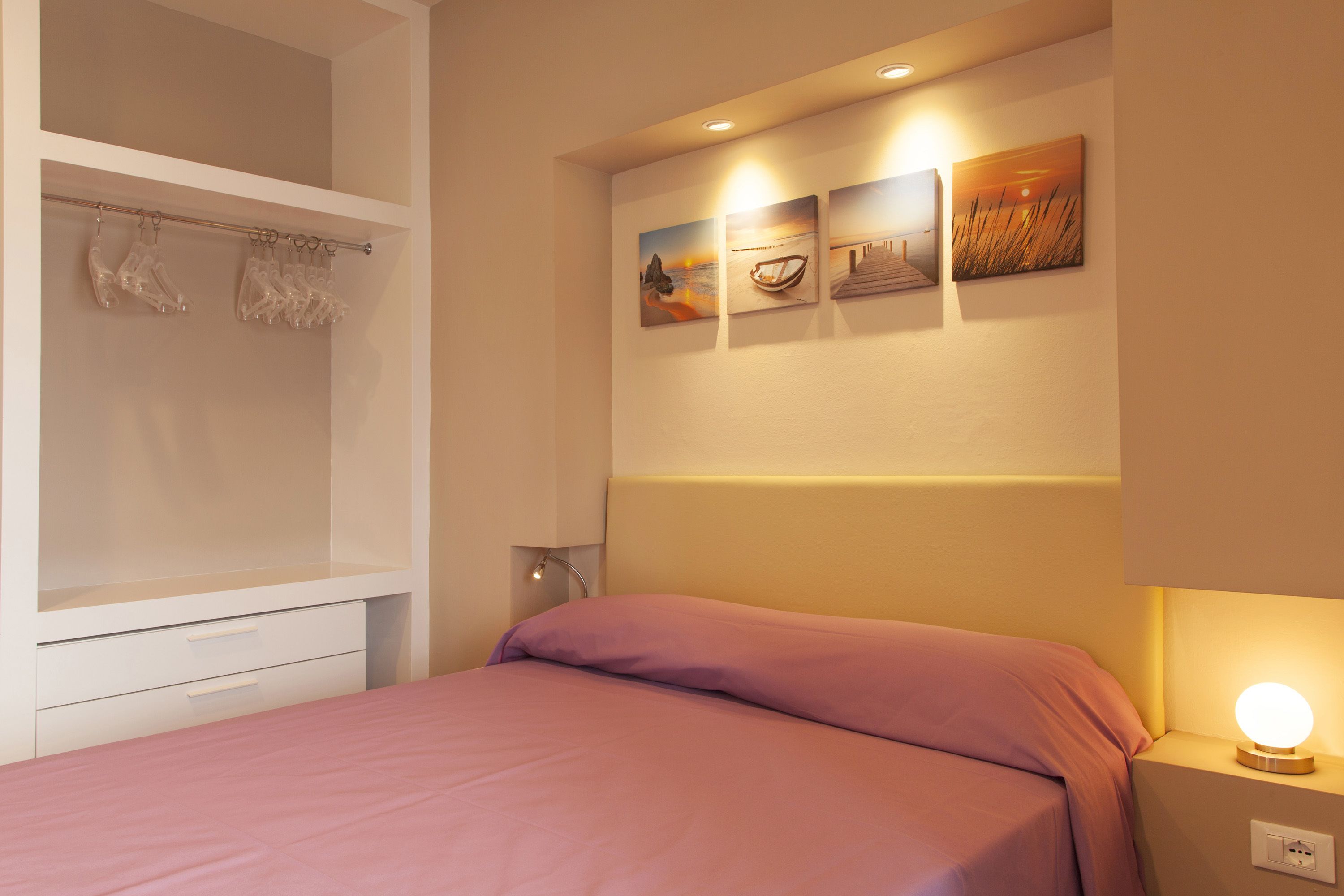 A bedroom with a bed, a nightstand, a wall-mounted shelf with decorative items, and a wall with three framed pictures.