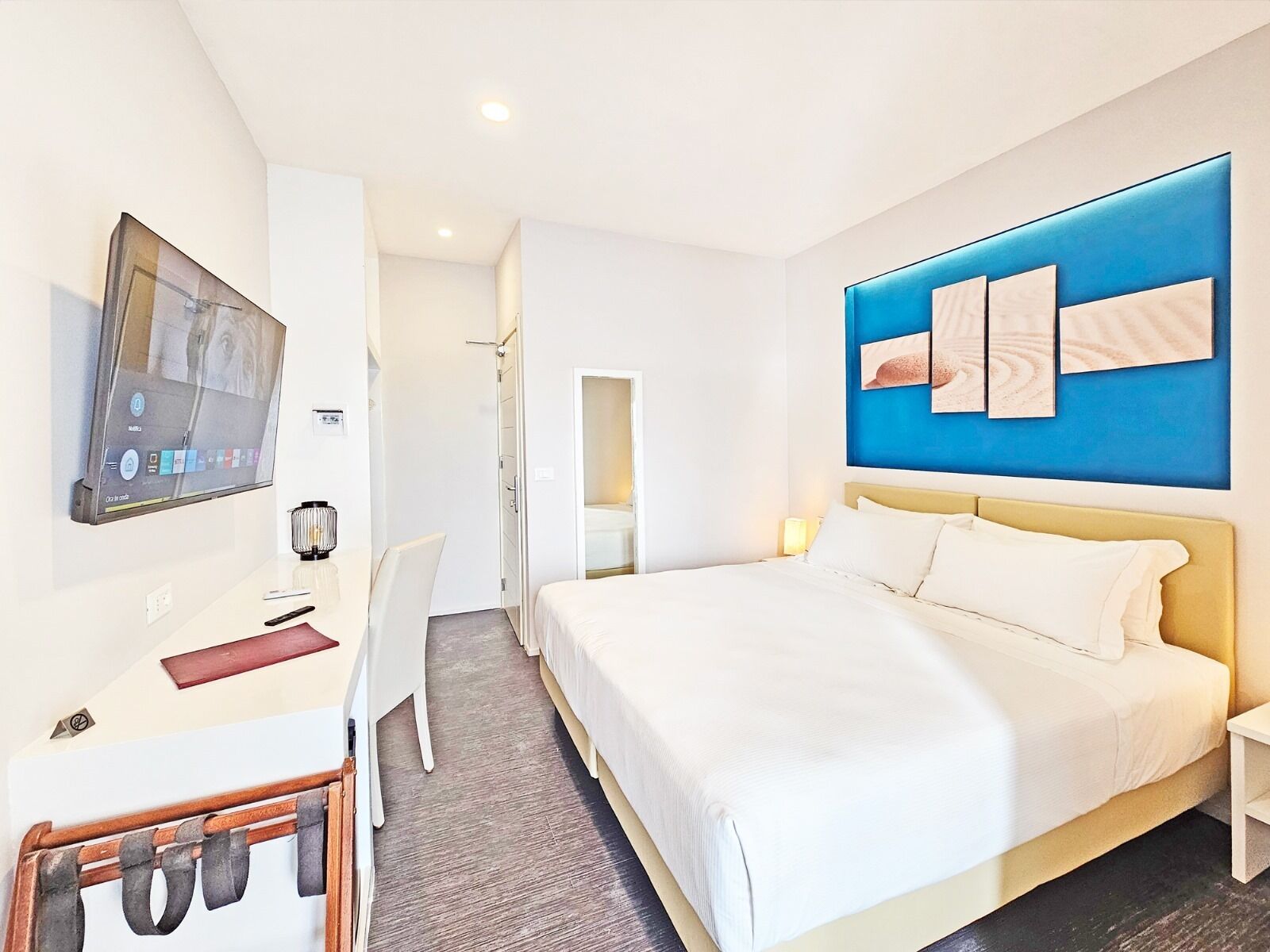 Ein modernes Hotelzimmer mit einem großen Bett, einem Schreibtisch mit Computer, einem an der Wand befestigten Fernseher und einer blauen Akzentwand.