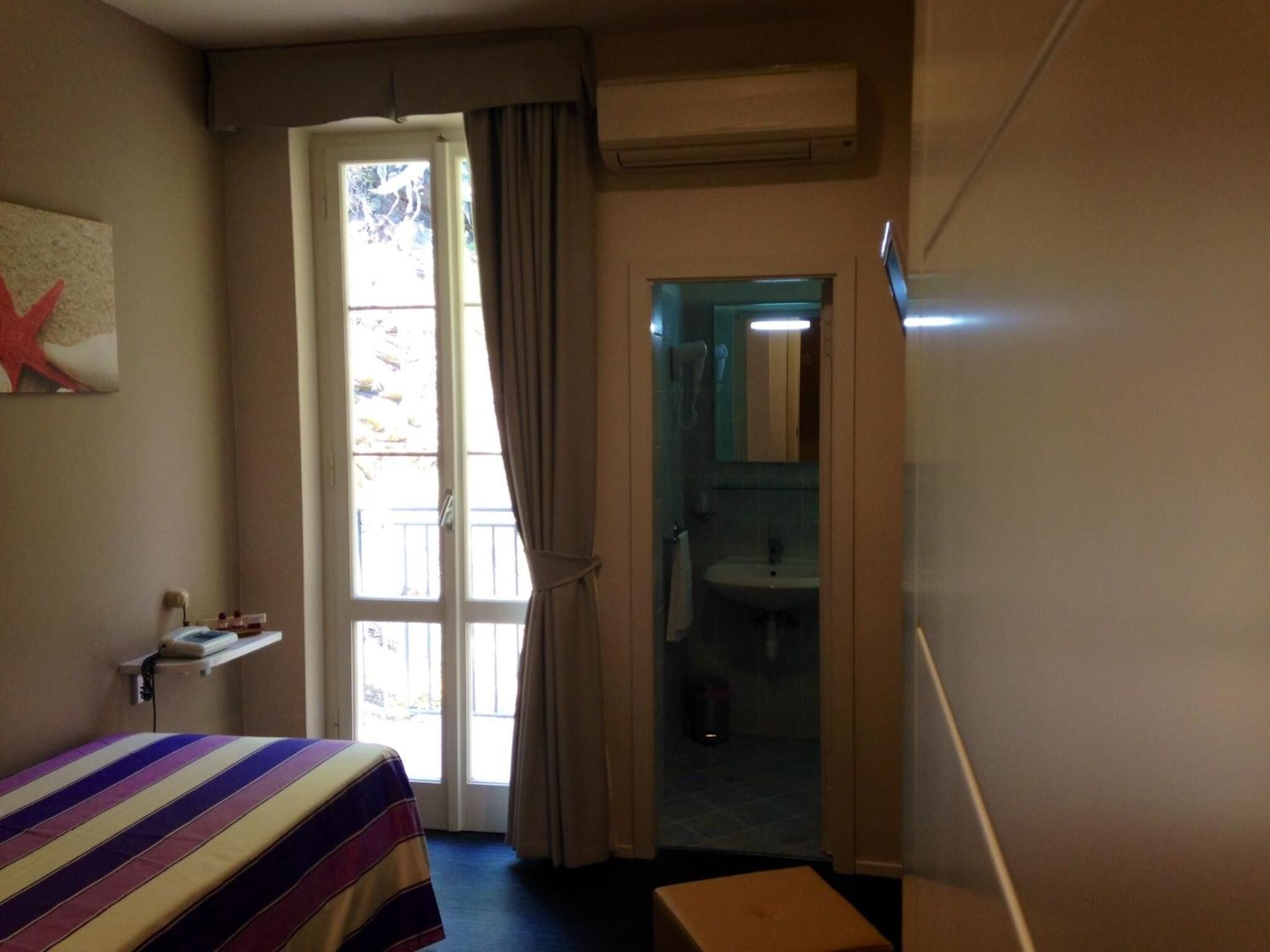 A hotel room with a bed, a small table with a lamp, a window with curtains, and a bathroom visible through an open door.