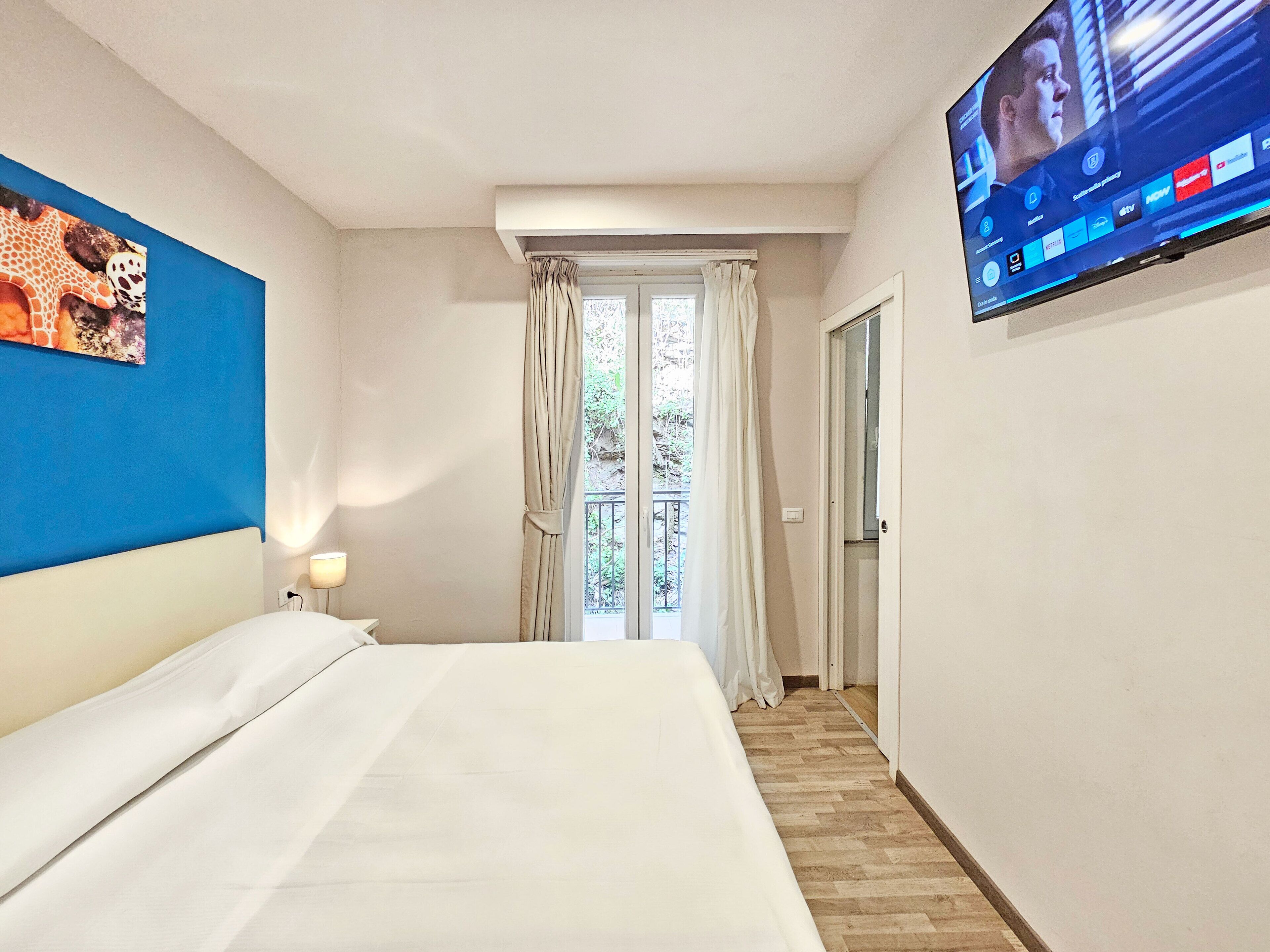 Ein Hotelzimmer mit Bett, einem an der Wand befestigten Fernseher, einem Fenster mit Vorhängen und Blick ins Freie.