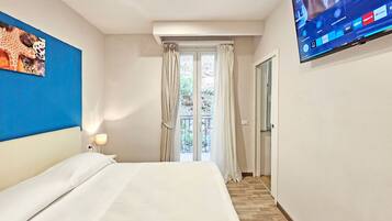 Ein Hotelzimmer mit Bett, einem an der Wand befestigten Fernseher, einem Fenster mit Vorhängen und Blick ins Freie.