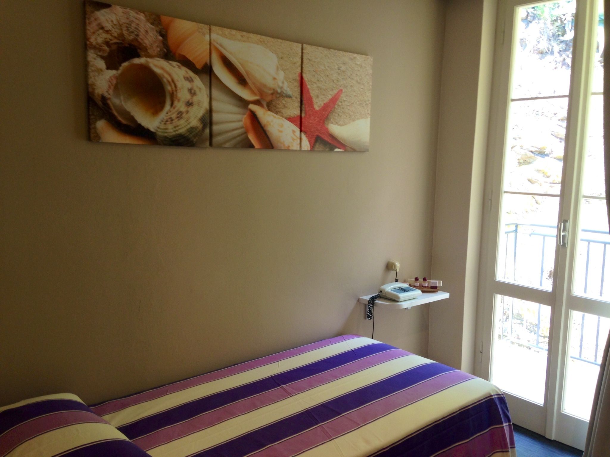 A room with a bed, a wall-mounted shelf with toiletries, and a large window with a view of the outdoors.
