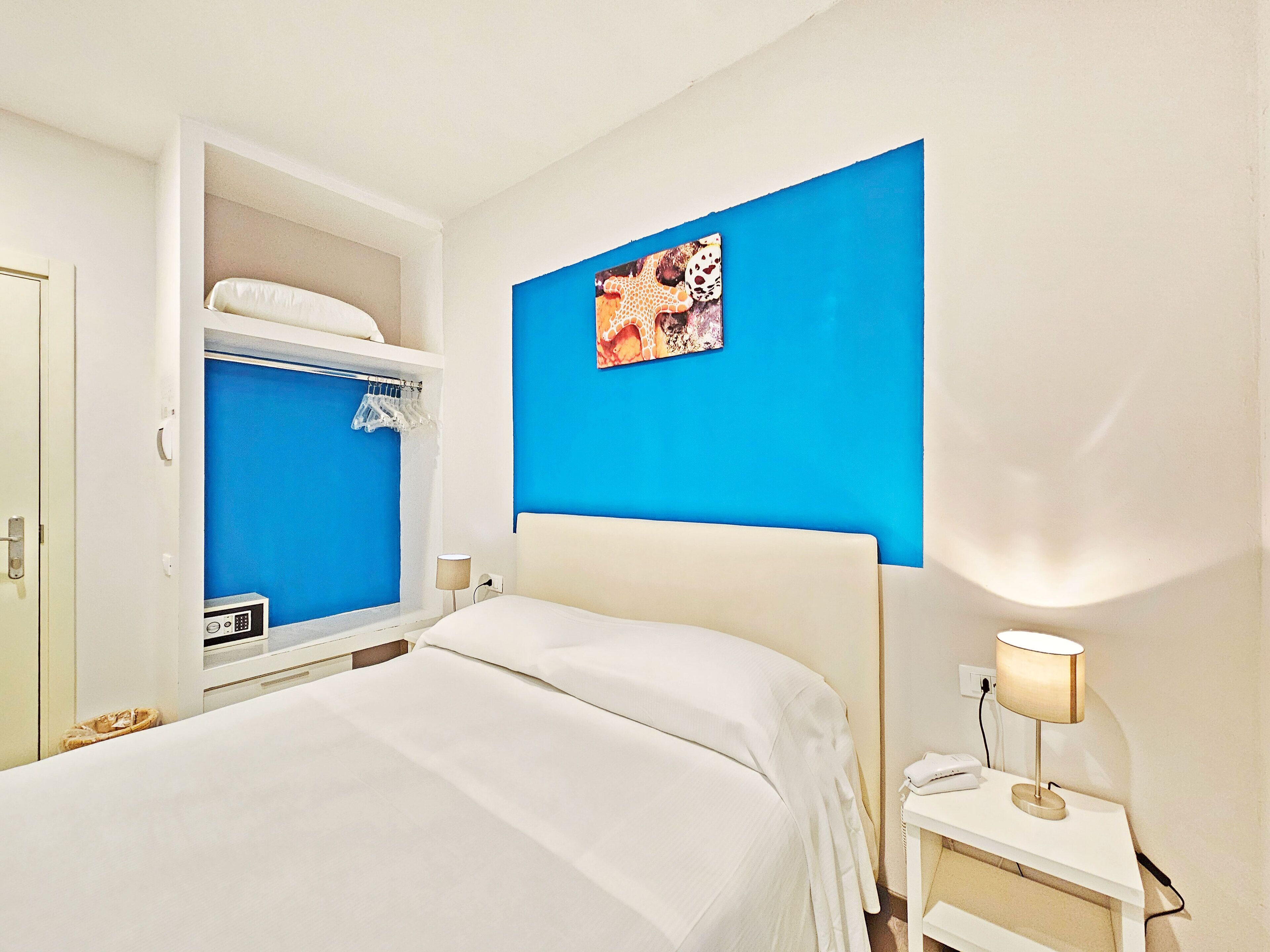 Una habitación de hotel con cama, mesita de noche, lámpara, armario y un cuadro en la pared.