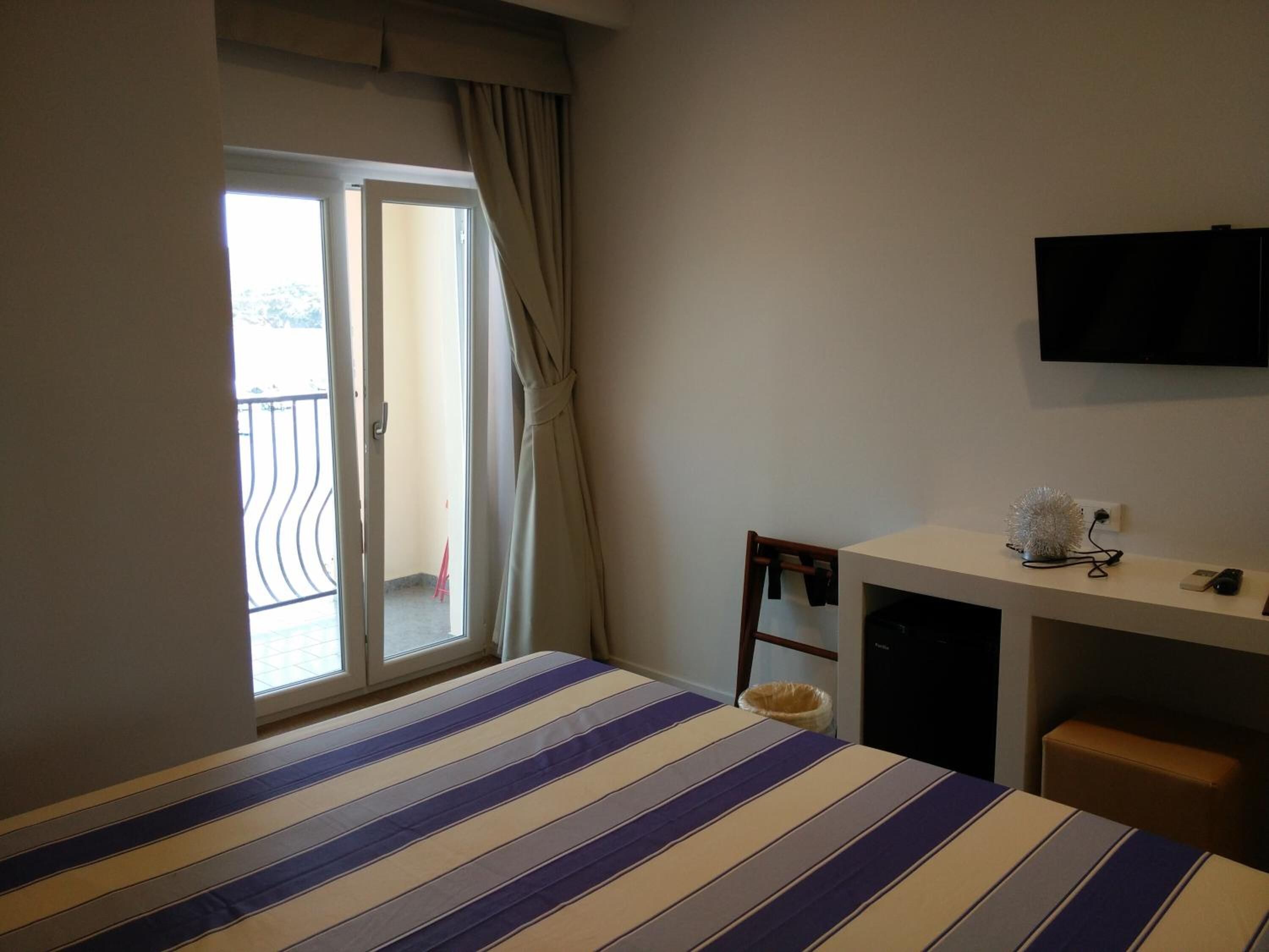 Habitación de hotel con cama, escritorio, silla, televisor y balcón con cortinas.