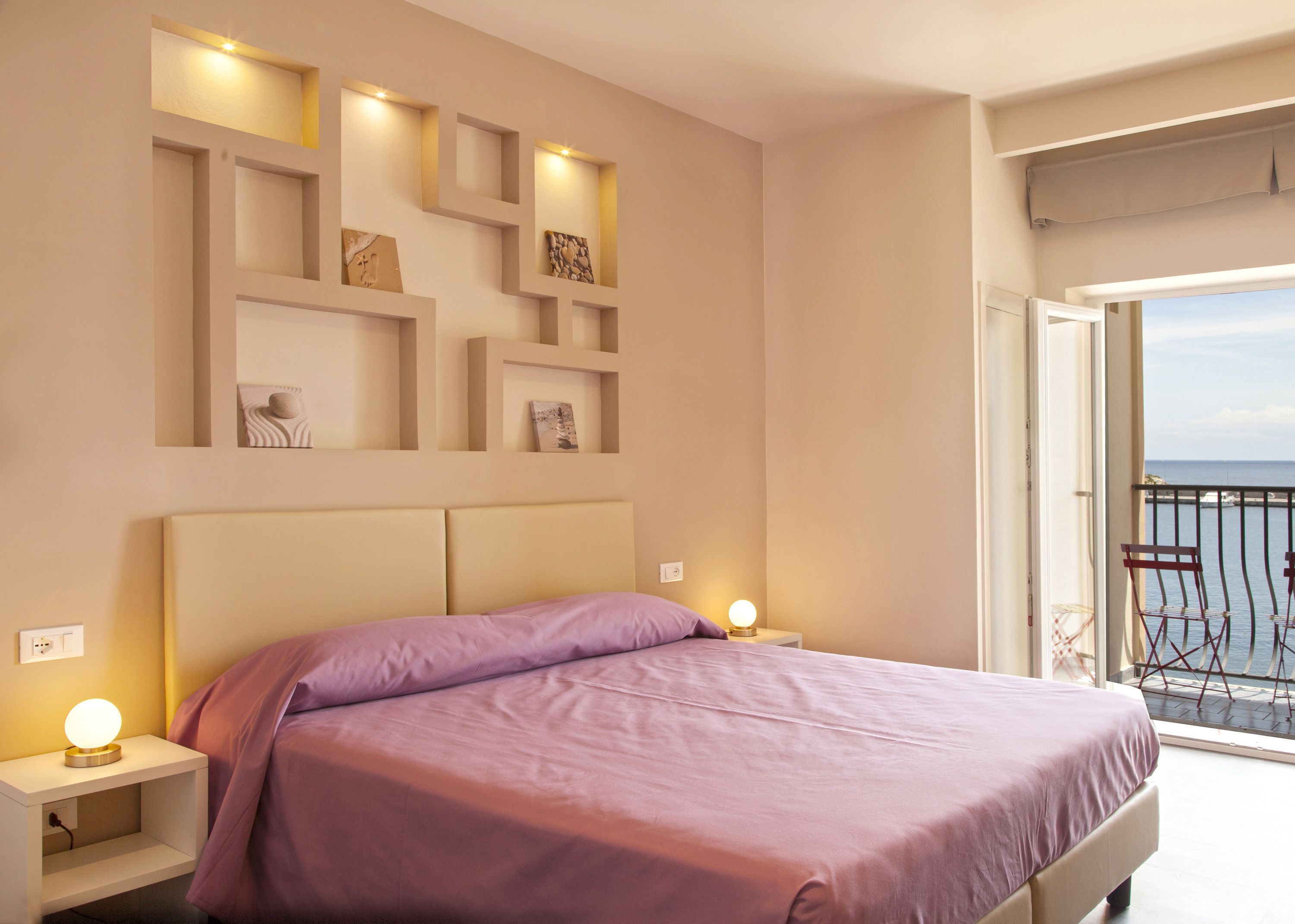 A bedroom with a bed, bedside lamps, and a balcony with a view of the sea.