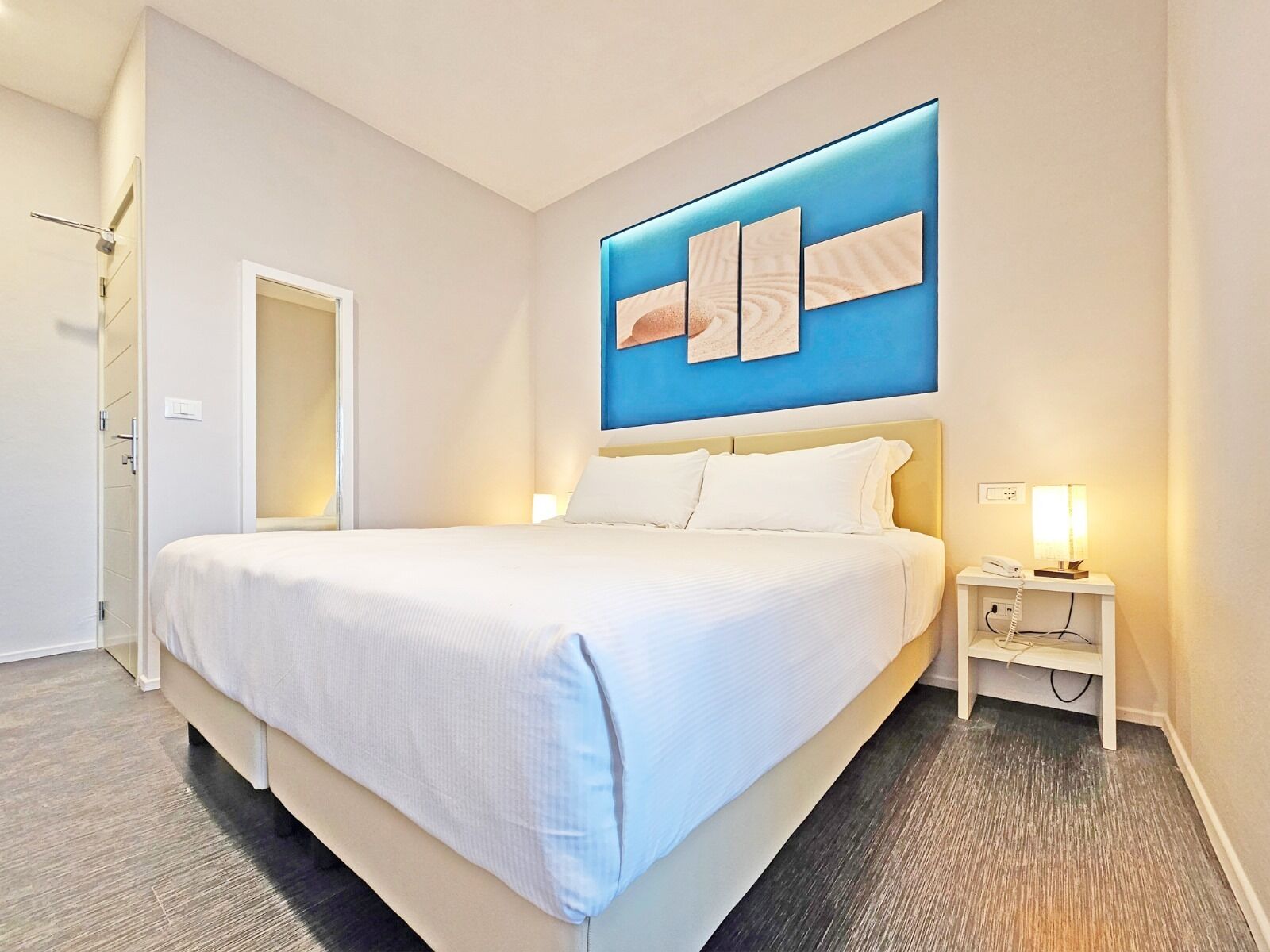 A modern hotel room with a large bed, a bedside table with a lamp, and a wall-mounted artwork.