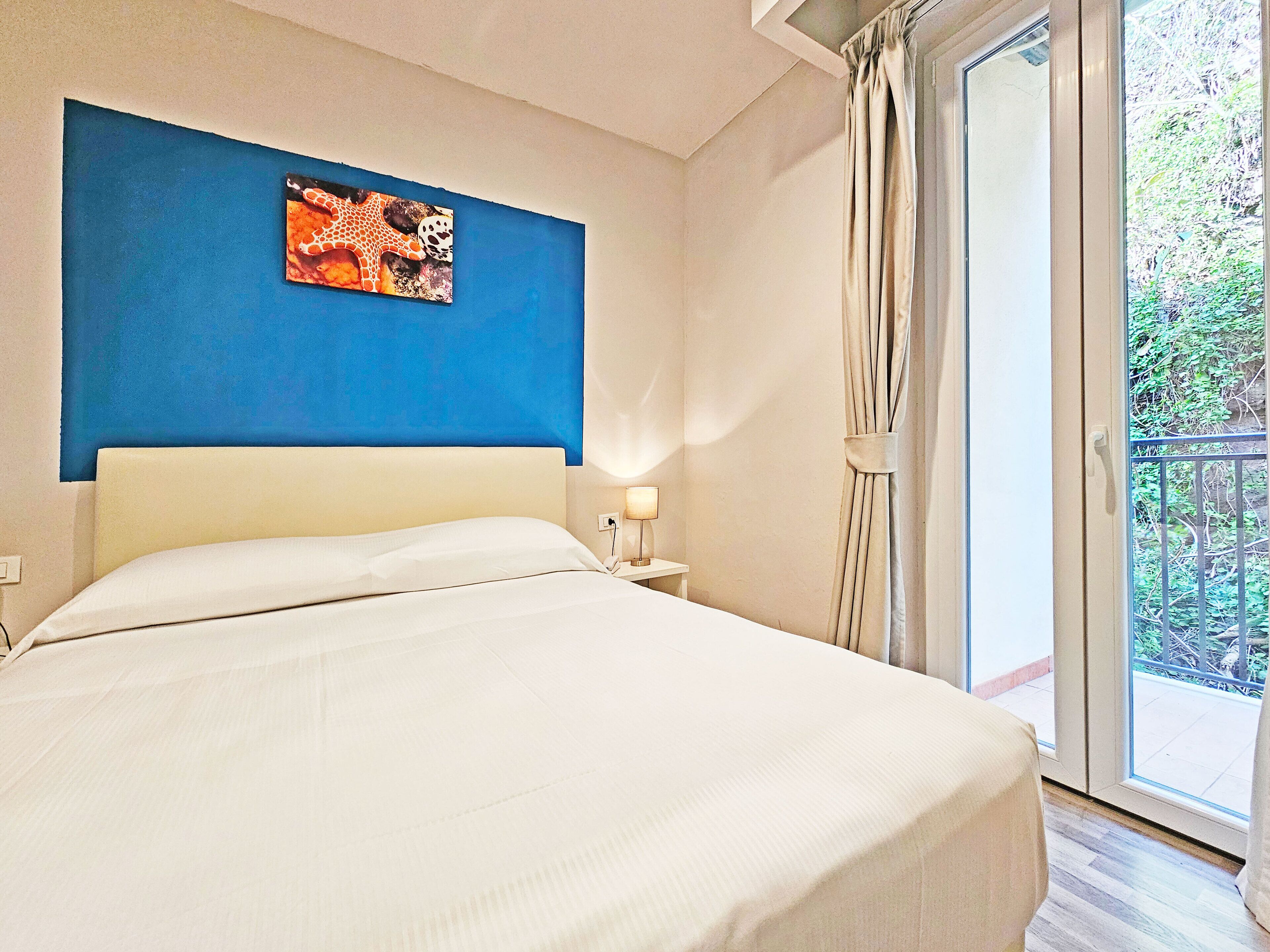 Ett hotellrum med en säng, ett nattduksbord, ett konstverk som hänger på väggen och utsikt över grönområden genom ett fönster.