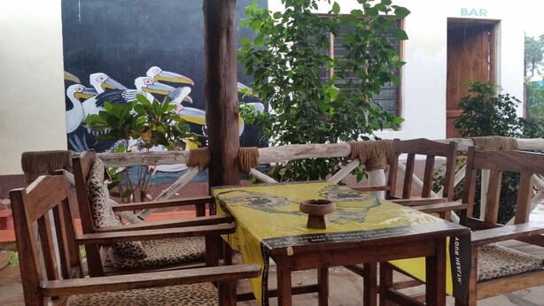 A wooden outdoor seating area with a yellow tablecloth, wooden chairs with leopard print cushions, and a mural of birds on the wall behind. - St. Gotthard Motel Bar & Restaurant (Malindi)