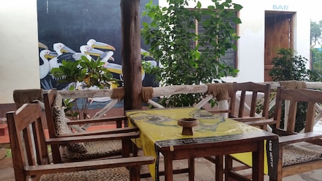 A wooden outdoor seating area with a yellow tablecloth, wooden chairs with leopard print cushions, and a mural of birds on the wall behind.