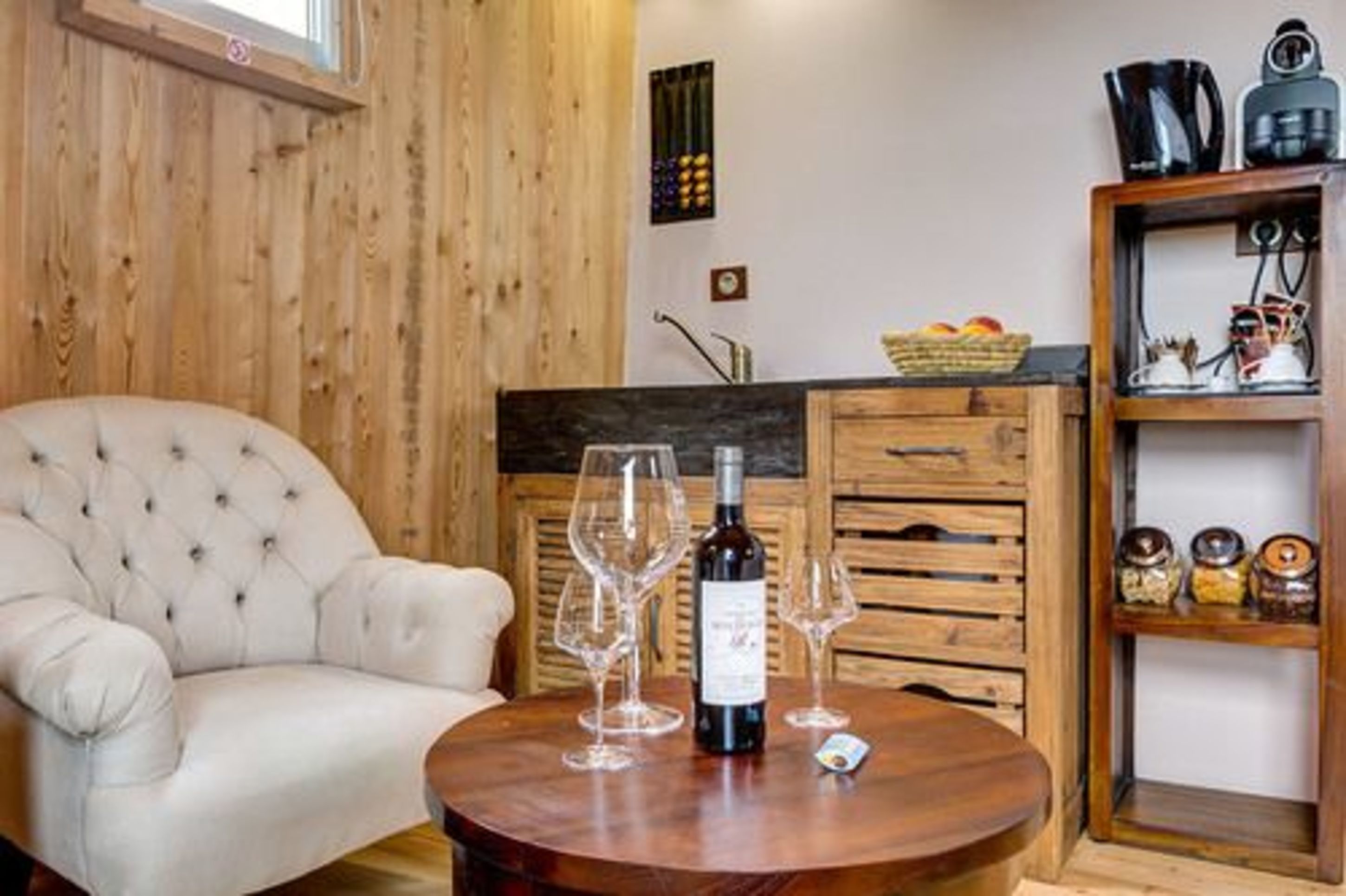 Una habitación acogedora con un sillón capitonado, una mesa de comedor de madera con copas de vino y una botella, y una zona de cocina con armarios de madera y una cesta de frutas.