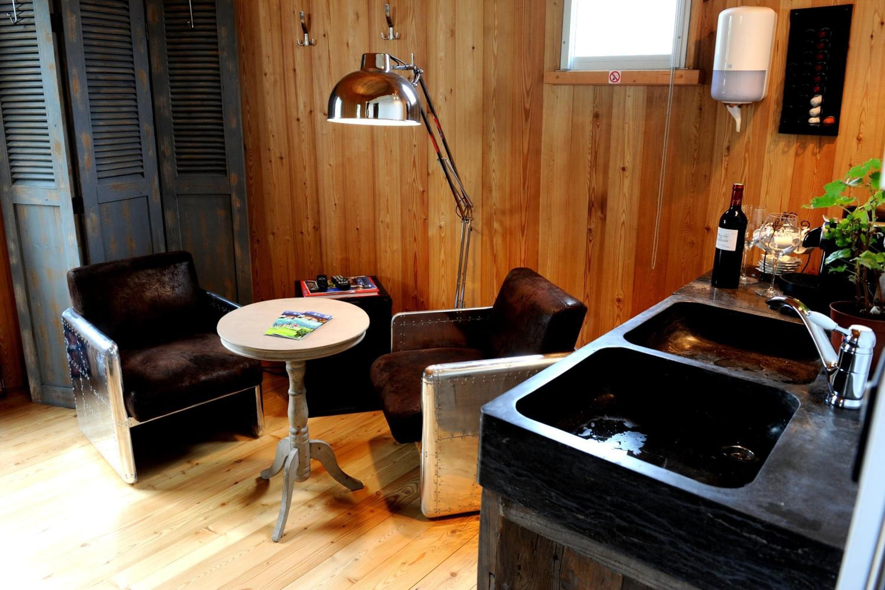 A cozy room with wooden walls, a round table, two leather chairs, a metal lamp, a sink, and a wine bottle.