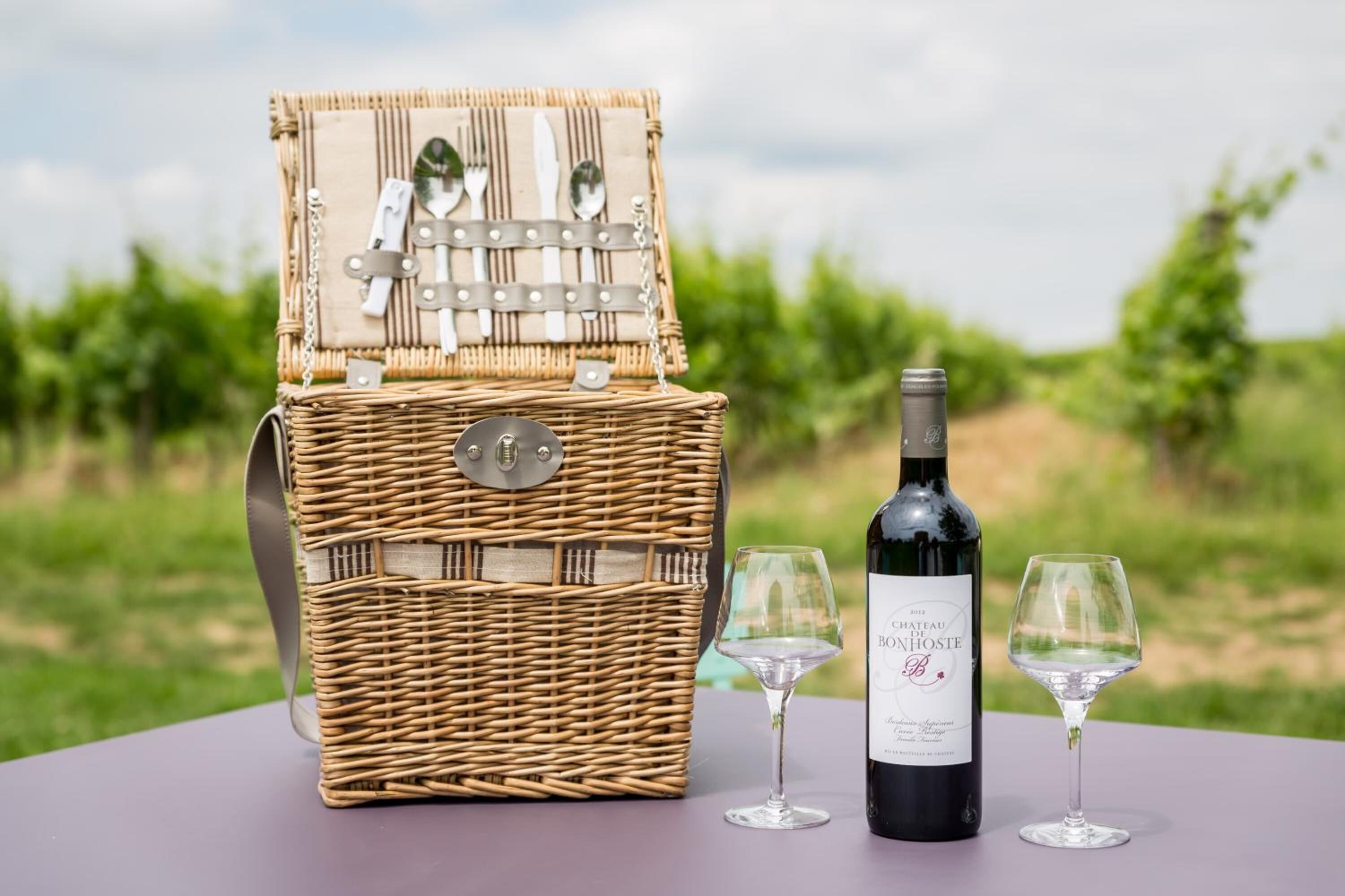A wicker picnic basket with a wine bottle and two glasses on a table with a vineyard in the background.