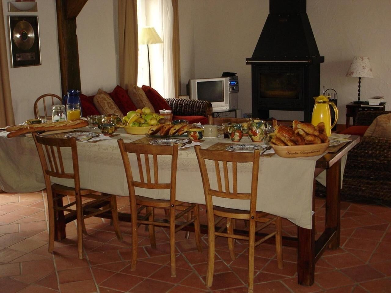 Un coin repas avec une table dressée pour quatre personnes, entourée de chaises en bois. Un téléviseur est fixé au mur, et on aperçoit un poêle noir au fond.