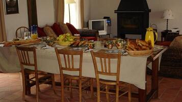 Un coin repas avec une table dressée pour quatre personnes, entourée de chaises en bois. Un téléviseur est fixé au mur, et on aperçoit un poêle noir au fond.