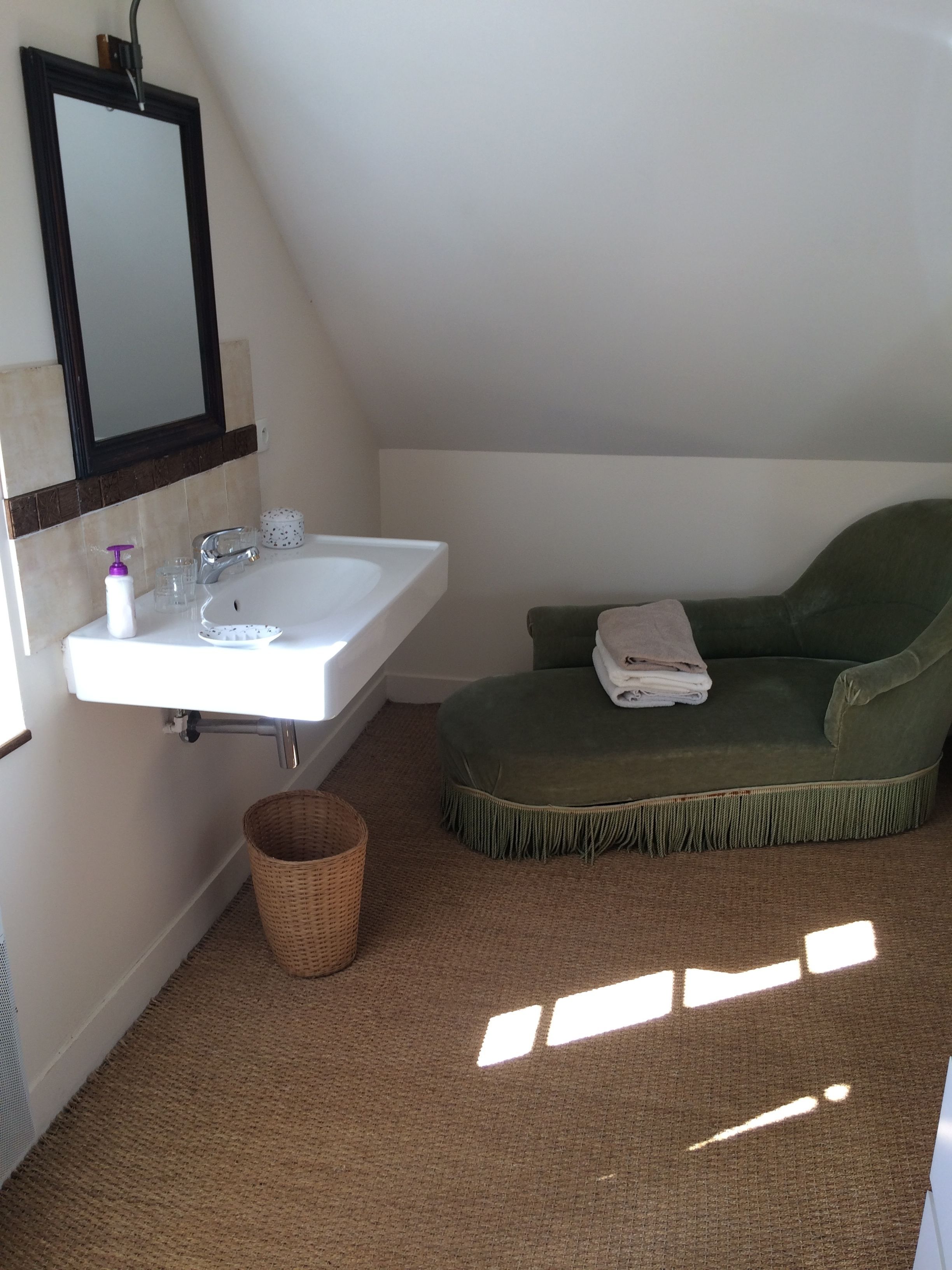 Un baño con lavamanos blanco, un espejo, un banco acolchado verde y una cesta de mimbre.