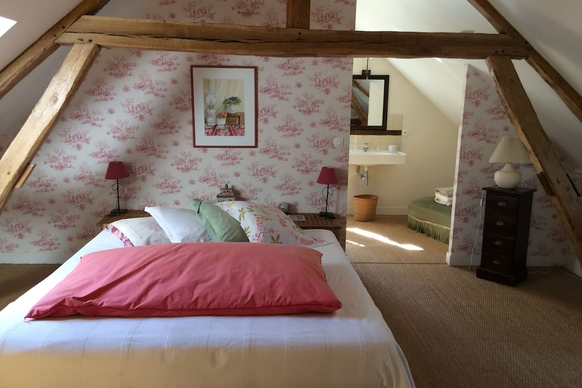 A bedroom with a sloped ceiling, floral wallpaper, a bed with pink and white bedding, two bedside lamps, a framed picture on the wall, and a small wooden nightstand.