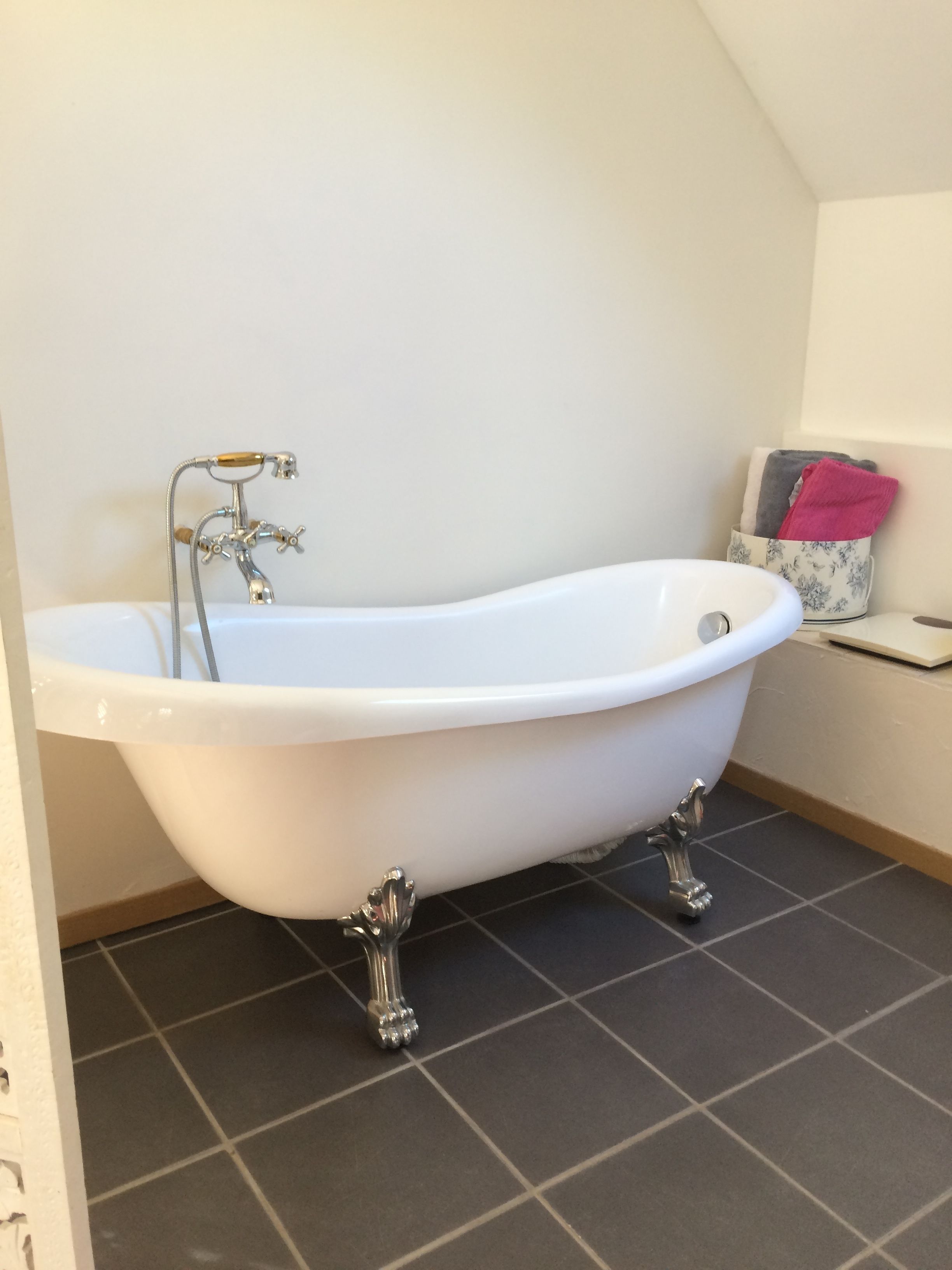 A white freestanding bathtub with a wooden handle, a chrome faucet, and a chrome base. There is a small shelf with towels and a decorative item. The floor is tiled with dark gray tiles.