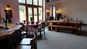 Un espace repas avec des tables et des bancs en bois, de grandes fenêtres avec des rideaux et des lampes fixées au mur.