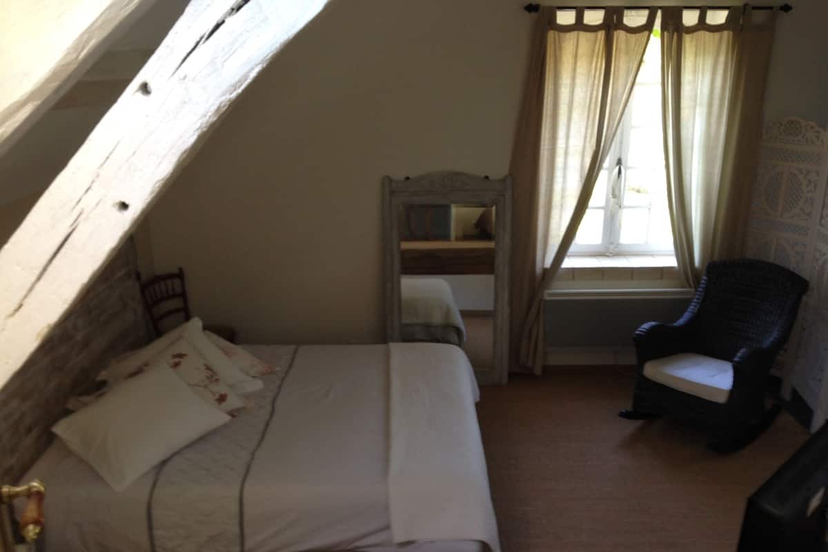 A bedroom with a bed, a chair, a window with curtains, and a mirror.