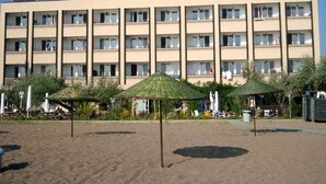 A multi-story building with balconies, a sandy ground, and two green umbrellas. - Dikili Sunset Club (Dikili)