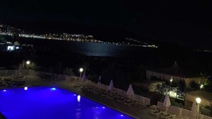 A nighttime view of a swimming pool with a blue-lit water feature, surrounded by lounge chairs and umbrellas, with a cityscape in the background. - Dikili Sunset Club (Dikili)