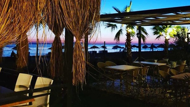 An outdoor seating area with tables and chairs, a thatched umbrella, and a view of the ocean at sunset. - Dikili Sunset Club (Dikili)