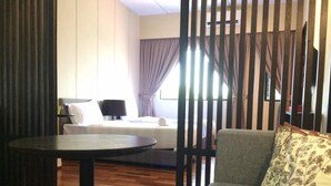 Quarto de hotel com piso de madeira, uma mesa redonda, um sofá com almofadas estampadas e uma janela com cortinas.