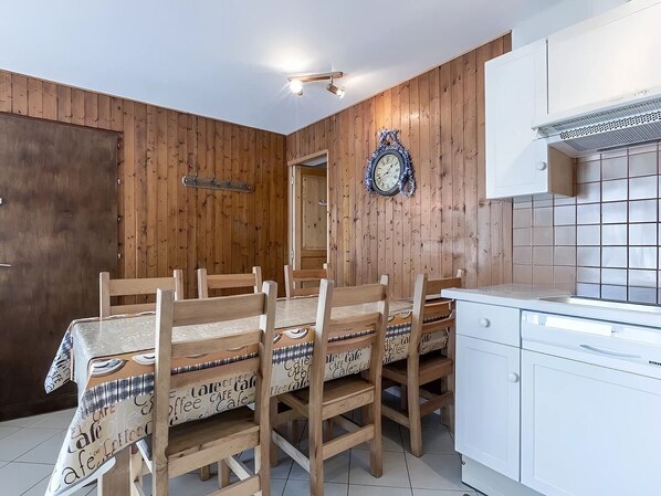 A dining area with a wooden table and chairs, a clock on the wall, and a kitchen with white cabinets.