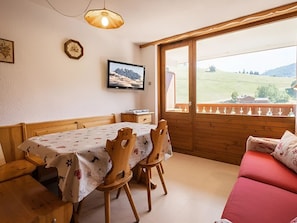 A dining area with a table, chairs, a television, and a view of a green landscape through a window.
