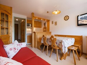 A cozy room with a red sofa, wooden dining table with chairs, a kitchen area, and a mounted TV. - 3* Apartment, 4-6 people, near slopes, pets allowed, garage (La Clusaz)