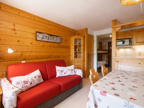 A cozy room with a red sofa, wooden walls, a cabinet, a microwave, and a table with a festive tablecloth. - 3* Apartment, 4-6 people, near slopes, pets allowed, garage (La Clusaz)