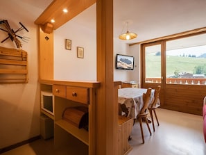 A room with a wooden dining table, chairs, a television, and a window with a view of a green landscape.