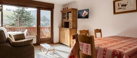 Una habitación con balcón, una mesa de comedor con sillas, una mecedora, un armario y un cuadro en la pared.