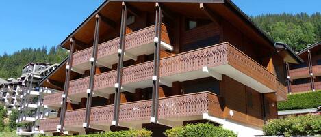 A multi-story wooden building with balconies, surrounded by greenery and a forested hillside.