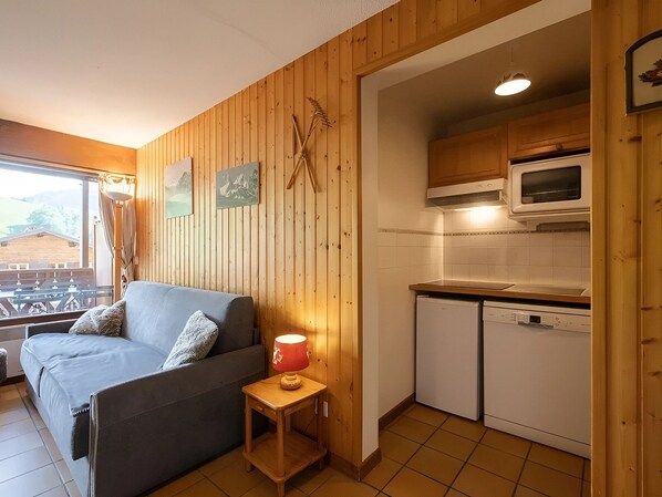 A cozy room with a sofa, a small table with a lamp, a kitchen area with a microwave and dishwasher, wooden walls, and a window with a view.