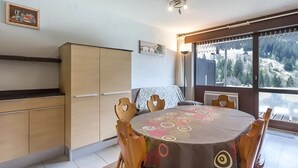 Ein Essbereich mit einem runden Tisch, bedeckt mit einer gemusterten Tischdecke, Holzstühle mit herzförmigen Ausschnitten, Blick auf eine verschneite Landschaft durch große Fenster und eine Küchenzeile mit Holzschränken.