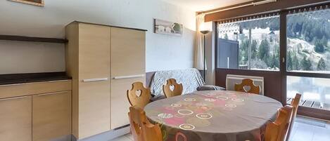 Ein Essbereich mit einem runden Tisch, bedeckt mit einer gemusterten Tischdecke, Holzstühle mit herzförmigen Ausschnitten, Blick auf eine verschneite Landschaft durch große Fenster und eine Küchenzeile mit Holzschränken.