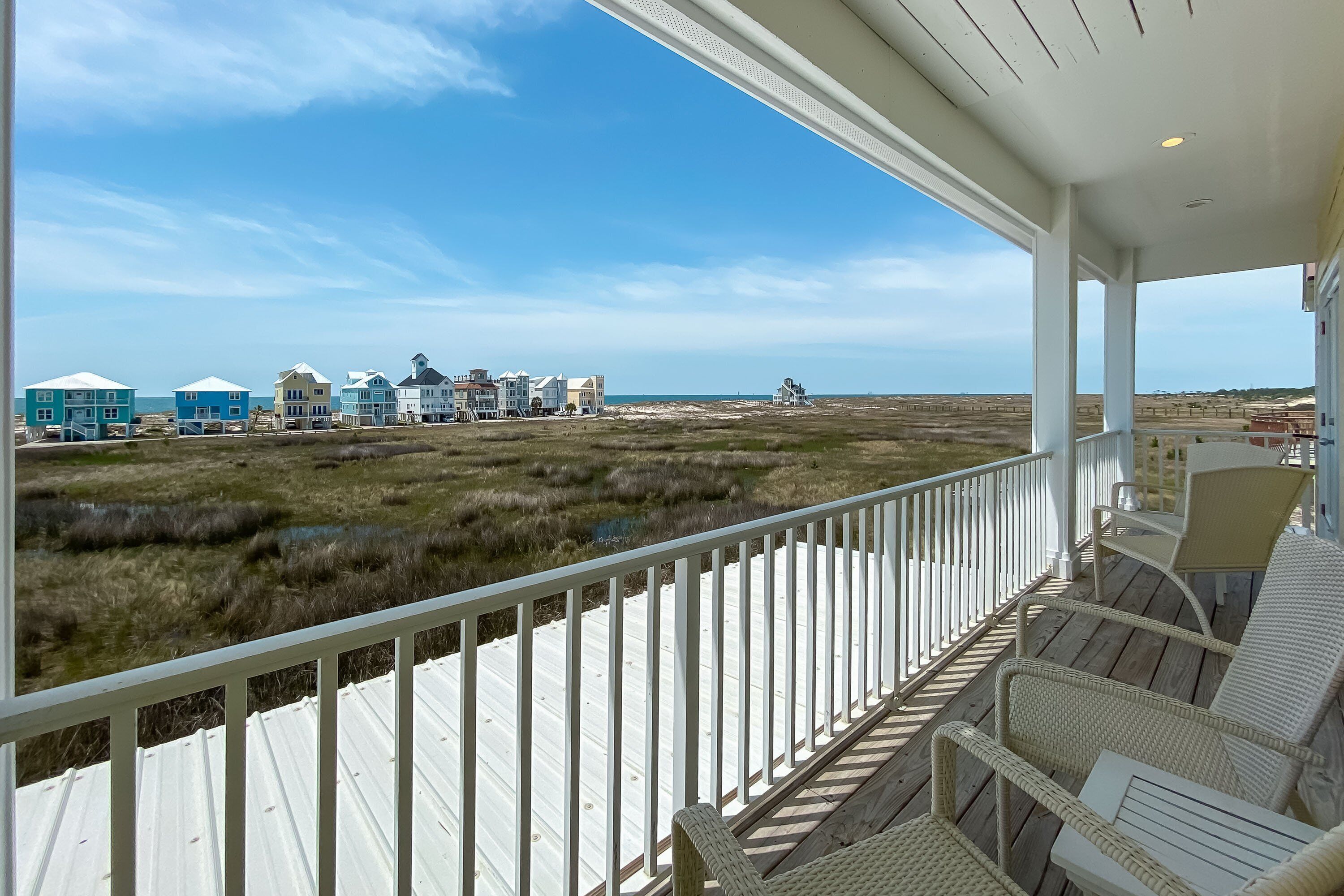 A wooden deck with wicker furniture overlooking a beach with houses in the background.