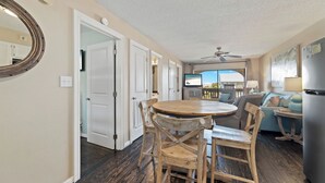 A dining area with a round wooden table and chairs, a ceiling fan, and a view of a balcony. - Come Stay At This Cozy Beach Condo! (Gulf Shores)