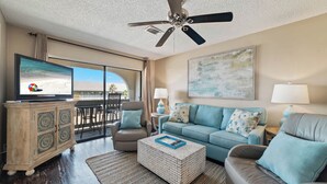 A spacious living room with a television, a ceiling fan, a sofa, a coffee table, and a cabinet. - Come Stay At This Cozy Beach Condo! (Gulf Shores)