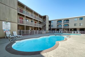 A pool area with a lifeguard sign, lounge chairs, and a multi-story building with balconies.