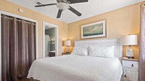 A bedroom with a bed, a ceiling fan, a nightstand with a lamp, and a painting on the wall. - Come Stay At This Cozy Beach Condo! (Gulf Shores)