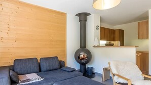 Un salon moderne avec un canapé d’angle gris, un poêle à bois noir, un fauteuil blanc moelleux et des murs recouverts de panneaux en bois.