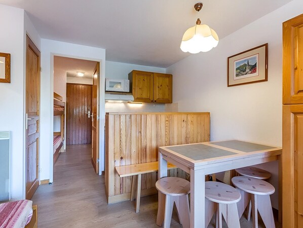 A compact kitchen with wooden cabinets, a microwave, a hanging light fixture, and a small dining area with stools.