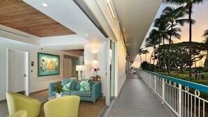 Un hall d’hôtel avec un plafond en bois, un canapé turquoise, des chaises jaunes et une vue sur les palmiers, visible à travers la rambarde d’un balcon.