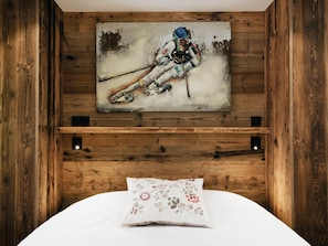 Ein Schlafzimmer mit einem hölzernen Kopfstück, einem Wandbild eines Skifahrers, einem weißen Bett mit einem Blumenkissen und einem Regal unterhalb des Bildes.